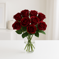 Ein realistischer Strauß aus zwölf dunkelroten künstlichen Rosen in einer klaren Glasvase auf weißem Tisch, vor hellem und minimalistischem Hintergrund.
