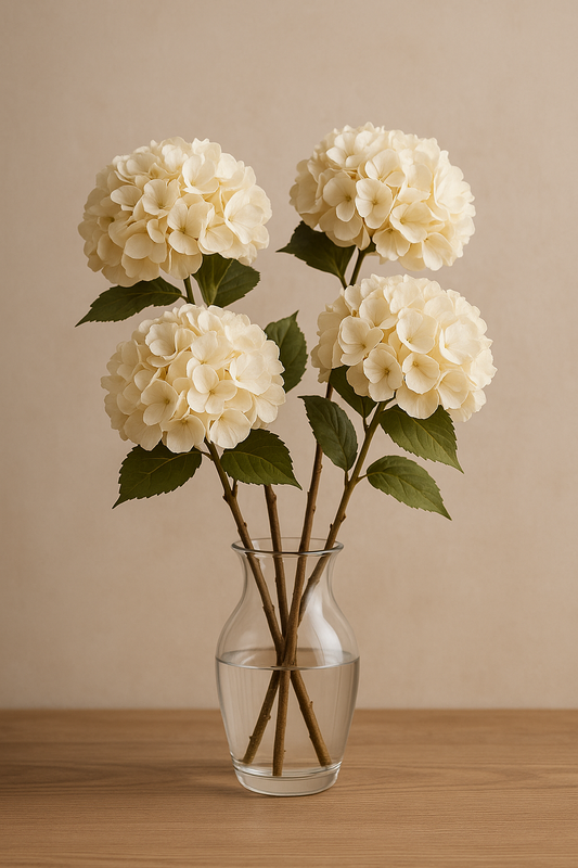 Vier cremefarbene künstliche Hortensien in einer Glasvase auf einem Holztisch vor einer beigen Wand – elegante Kunstblumen-Deko für Wohnzimmer, Hochzeit oder Tischgestaltung.